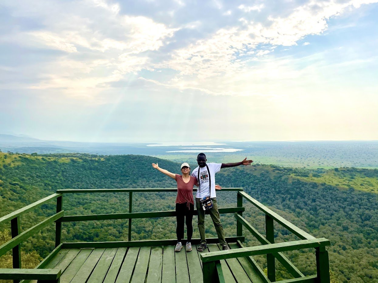 Two people standing on a green wooden platform with a vast green forest and lake in the background under a partly cloudy sky, with their arms open wide.