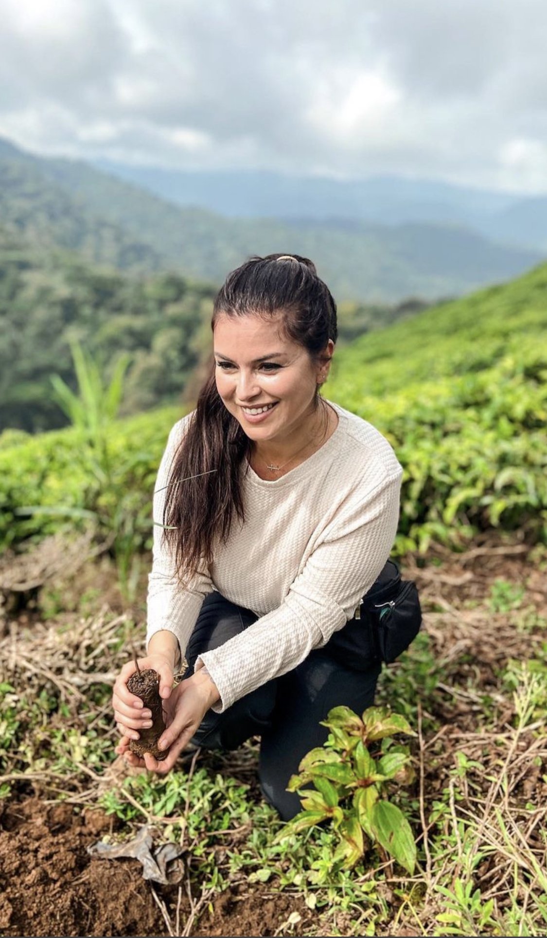 A woman kneeling in a field of crops, smiling while holding a small young plant with soil on its roots, with green hills and mountains in the background under cloudy skies.