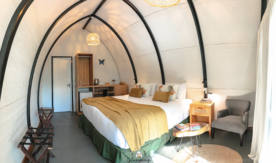 Cozy hotel tent room with a large bed, wooden side table, armchair, small table with books, wardrobe, and butterfly wall decoration.