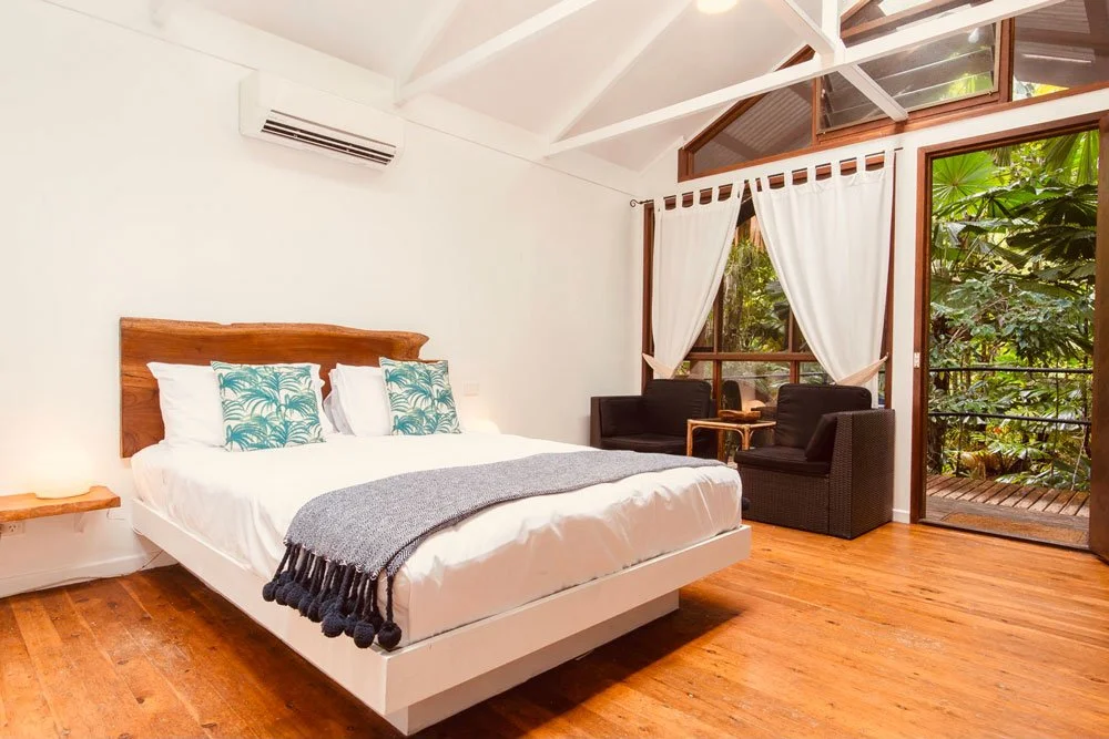 A bedroom with a large bed, black armchairs near large windows with white curtains, and a view of lush green trees outside.
