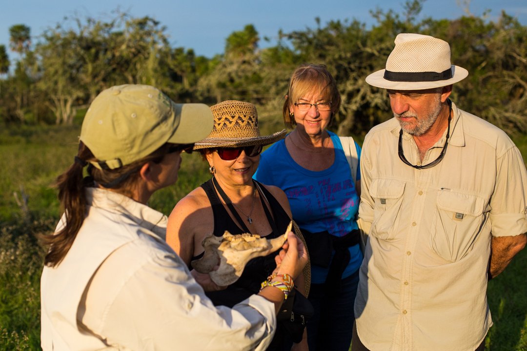 A tour guide showing a bone to a group of four tourists outdoors during daytime, with trees and blue sky in the background.