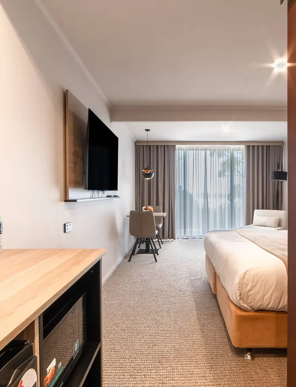 Hotel room with a bed, television, small dining table with chairs, and sliding glass doors with curtains overlooking an outdoor view.