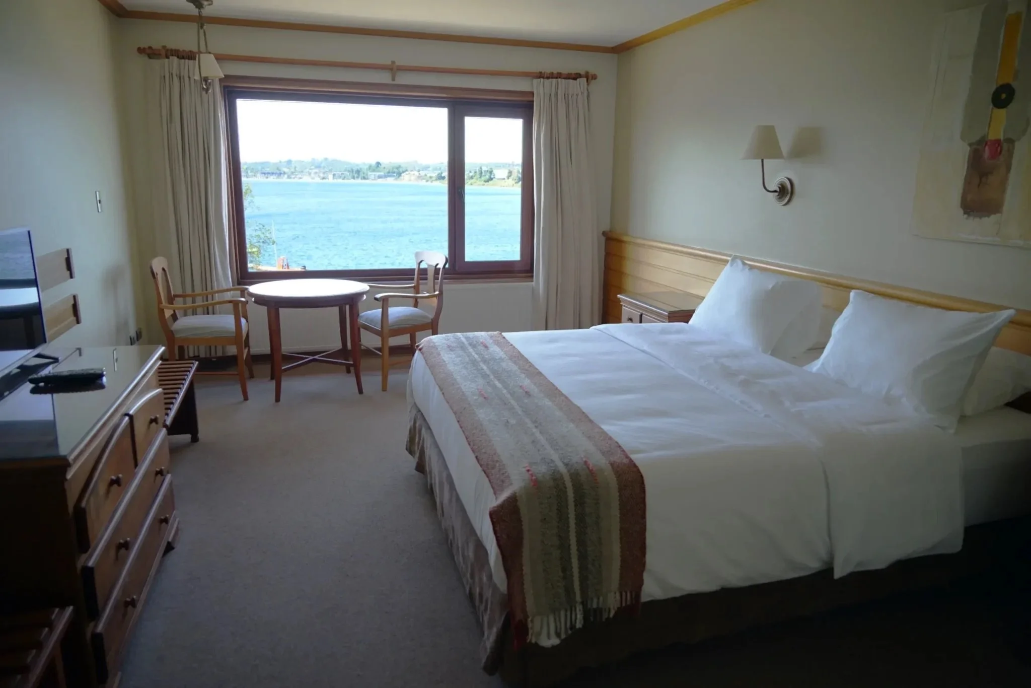 Hotel room with a large bed, a wooden headboard, two pillows, and a striped blanket. There is a window showing a view of a lake outside. The room has a small round table with two chairs near the window, a wooden dresser with a TV, and a wall-mounted light fixture.