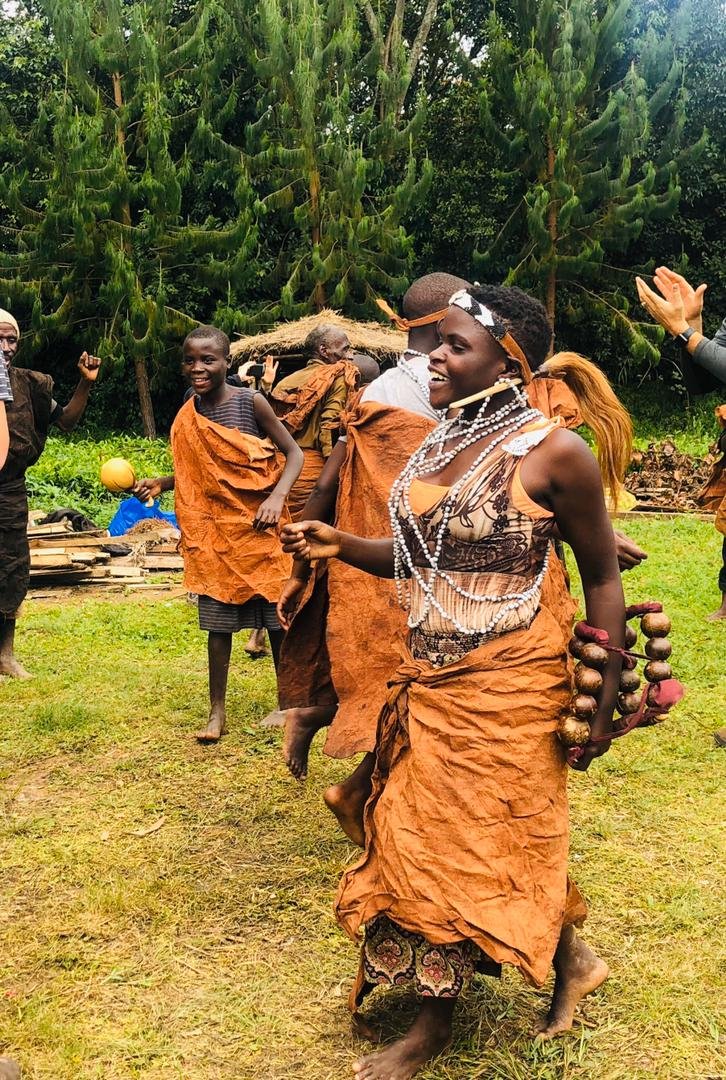 People dancing outdoors in traditional attire, with lush green trees in the background.