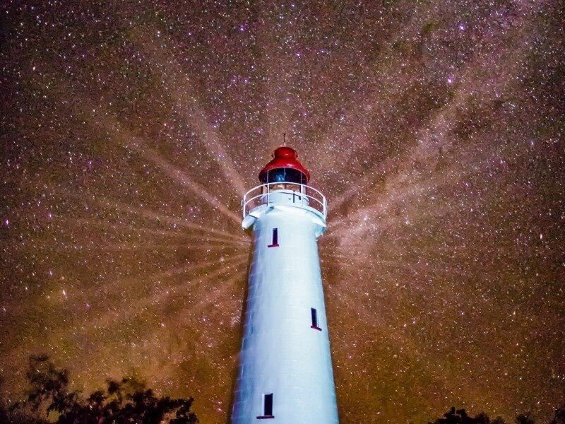 Lighthouse illuminated at night with the Milky Way galaxy and stars in the background.