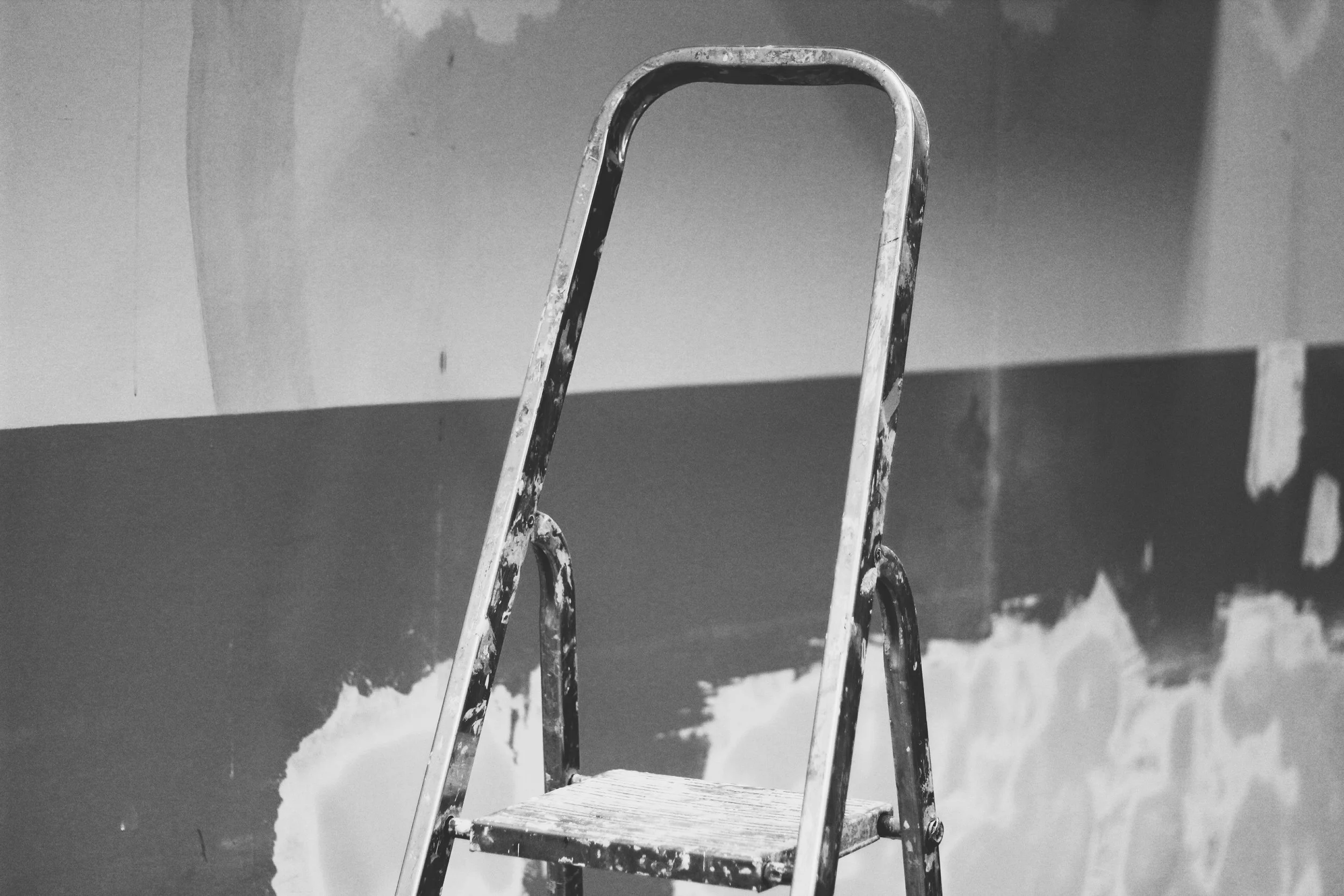 Black and white photo of a paint-splattered step ladder in front of a partially painted wall.