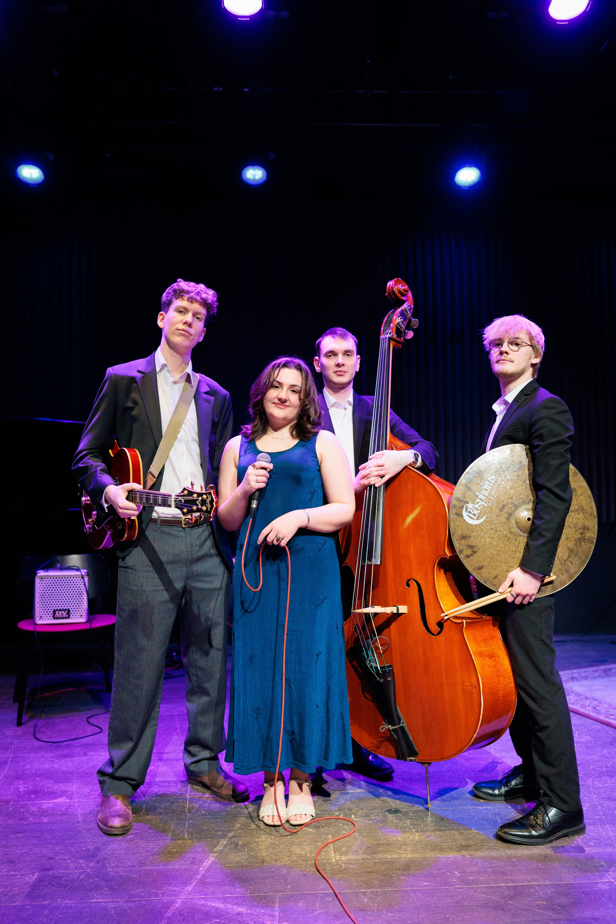 A musical quartet on stage: a guitarist, a vocalist with a microphone, a double bassist, and a drummer holding cymbals, under stage lights.