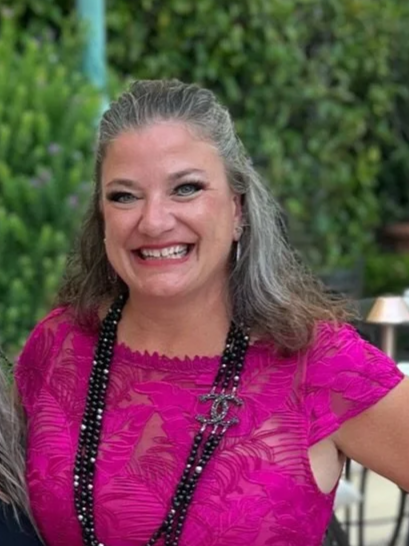 A woman with gray hair smiling, wearing a bright pink dress with a floral pattern, layered necklaces, and a ring on her right hand, standing outdoors against a green background.