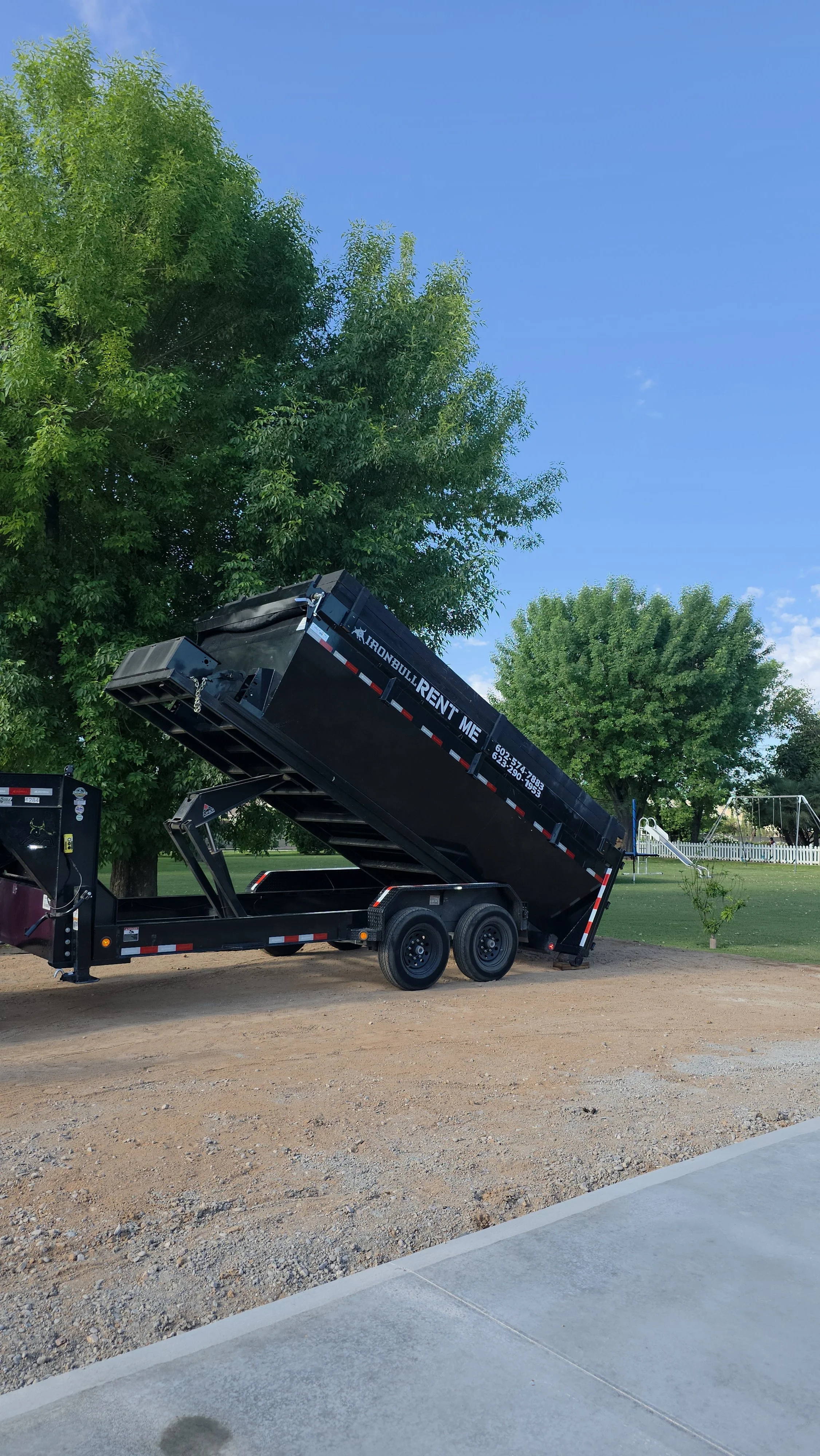Black dump trailer raised at an angle, parked on a dirt area with trees and playground in the background. Commercial/Residential 
Rental in Phoenix, AZ