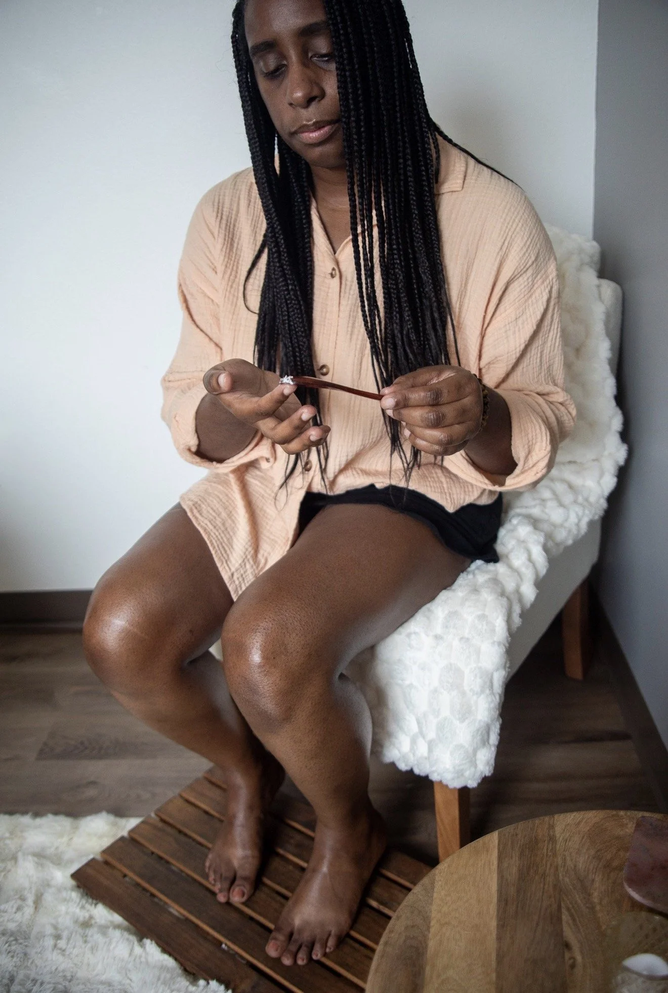 A woman with braided hair sitting on a white textured chair, looking at her phone, wearing a peach-colored shirt and black shorts, with her feet on a wooden bath mat on a hardwood floor.