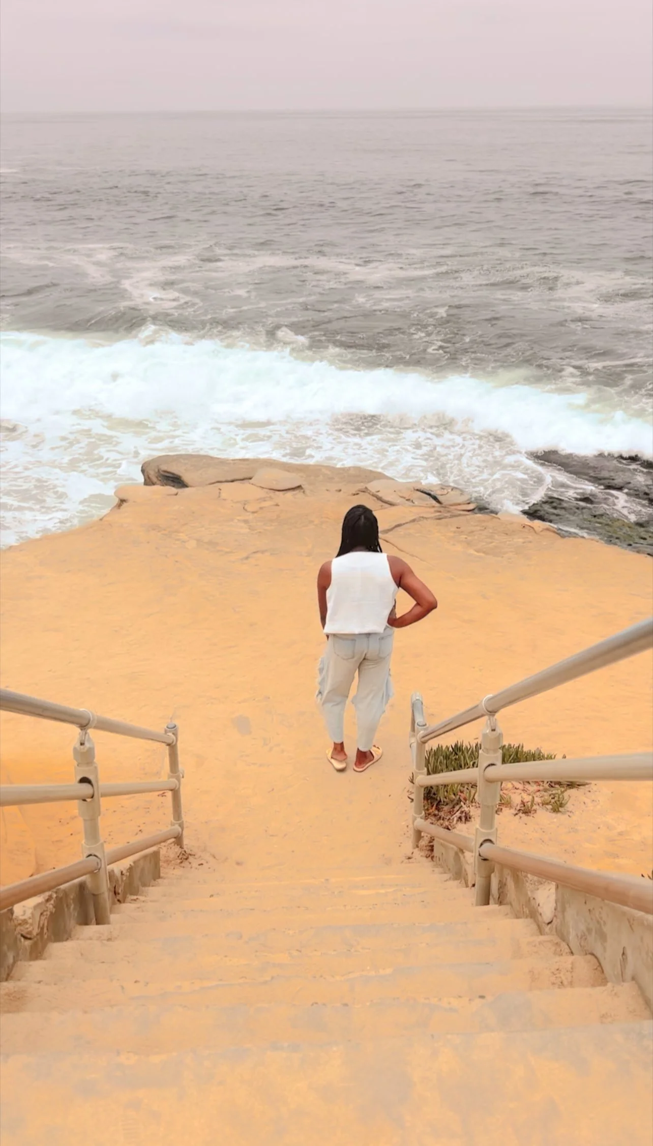 A woman standing at the top of a staircase leading down to a sandy beach by the ocean, with waves crashing against the shore.