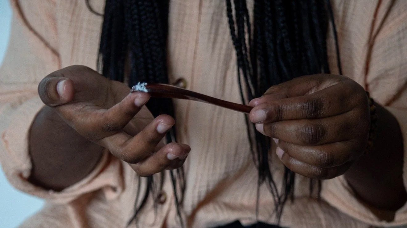 A person with dark skin holding a brown spoon with white powder in it, wearing a light peach long-sleeve shirt and black braids.