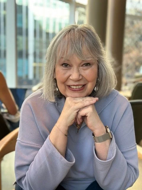 A smiling elderly woman with gray hair and a light purple sweater sitting indoors, resting her chin on her hands, in front of a window with natural light.