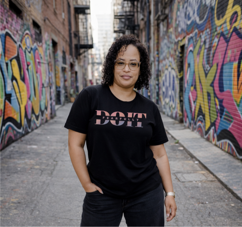 A woman with curly hair and glasses standing in a graffiti-covered alley, wearing a black T-shirt with pink and white text.