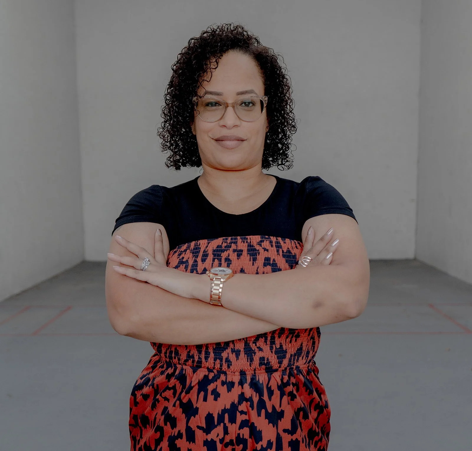 A woman with curly hair and glasses standing with her arms crossed in an all-white room.