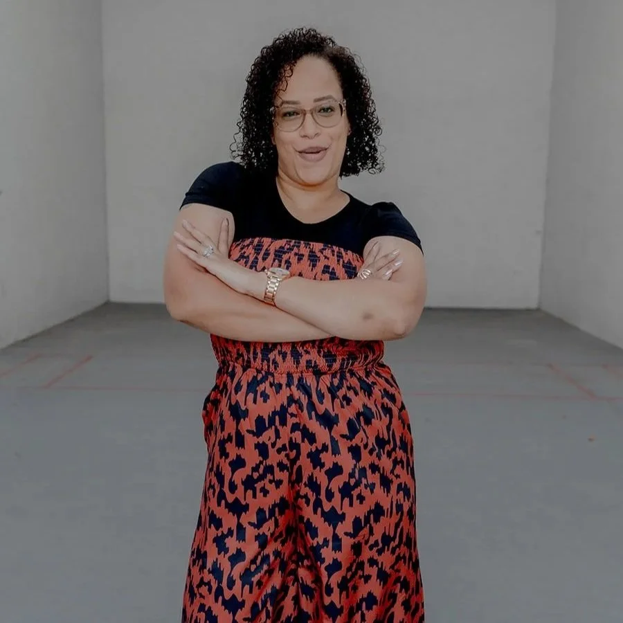 A woman with curly hair, glasses, and light skin, standing with her arms crossed, smiling, against a plain gray background.