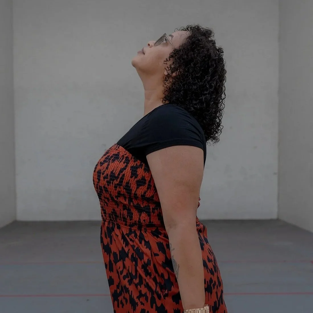 A woman with curly hair wearing glasses, a black and red patterned dress, standing against a plain gray wall, looking upward.