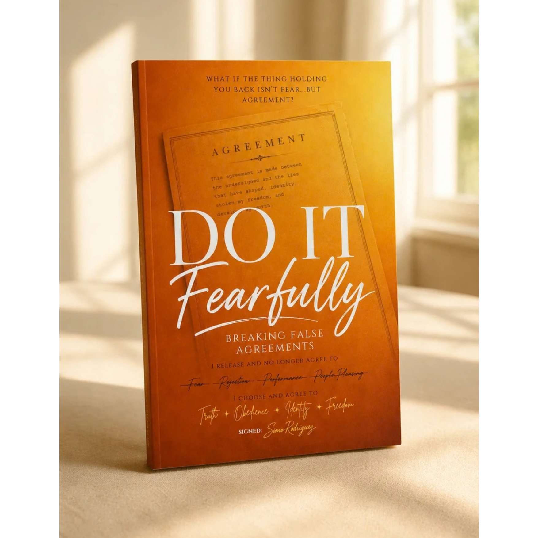 Close-up of an orange book titled "Do It Fearfully" with a subtitle "Breaking False Agreements" and a signed note at the bottom, placed on a surface with a blurred background of a window and cream-colored curtains.