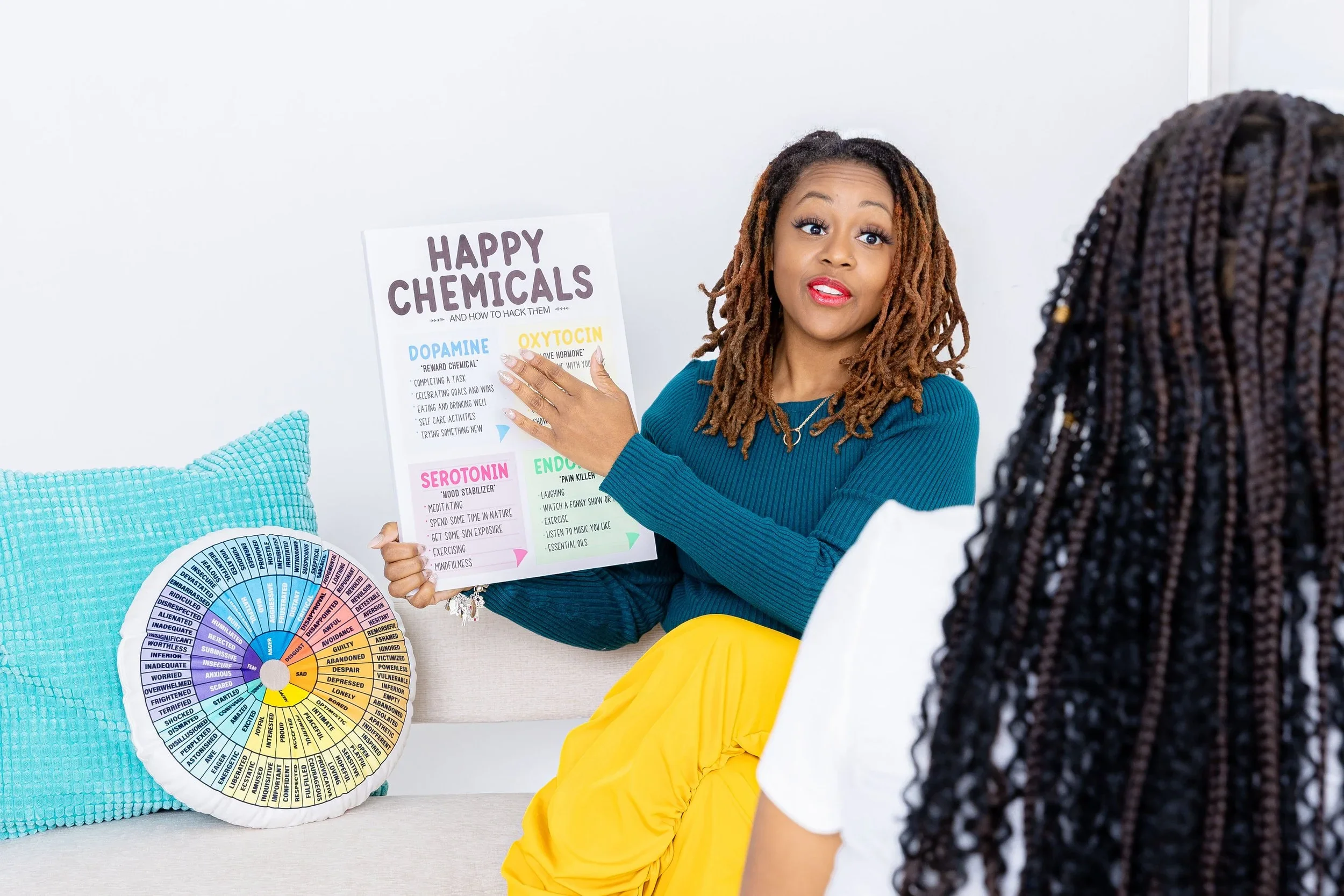 Woman giving a presentation about happiness chemicals to another woman, pointing to a poster with colorful text on a white wall.