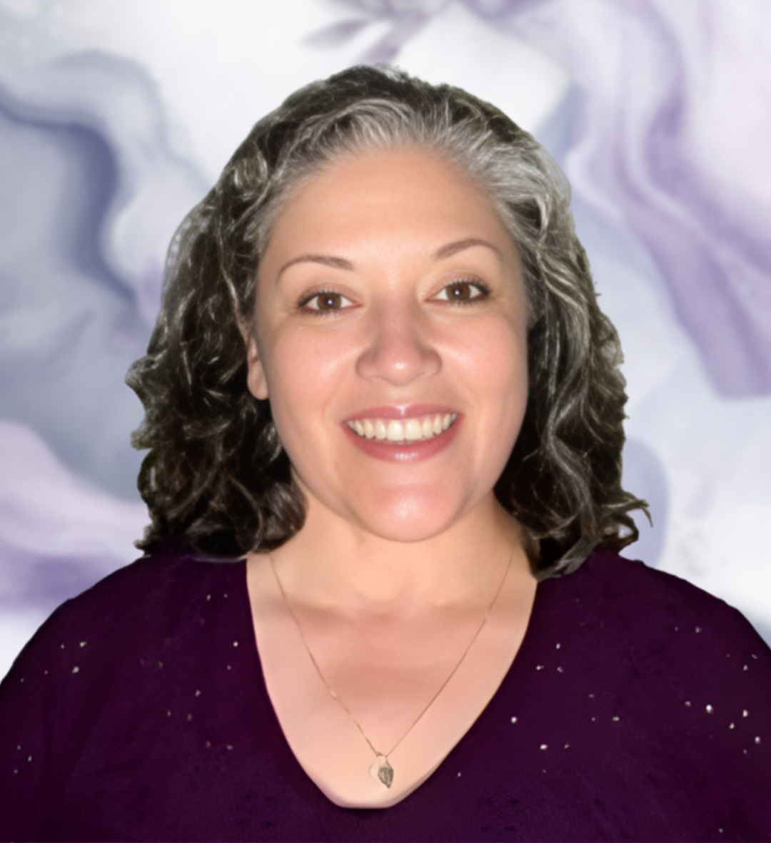 Portrait of a woman with curly gray hair, smiling, wearing a purple top with small sparkles and a necklace with a teardrop-shaped pendant, with a blurred abstract background.