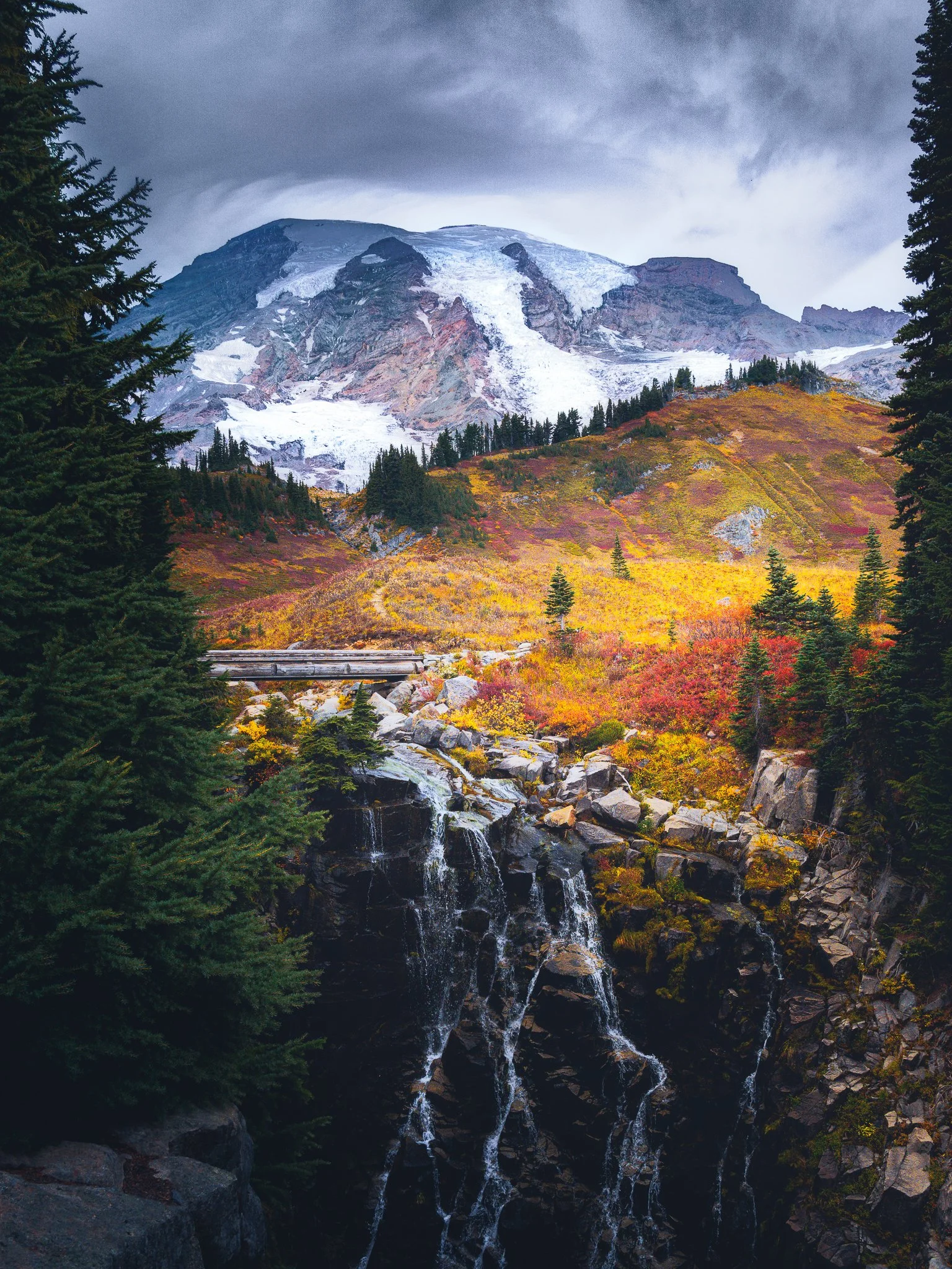 A scenic mountain landscape with a snow-capped peak, colorful autumn foliage, evergreen trees, and a waterfall.