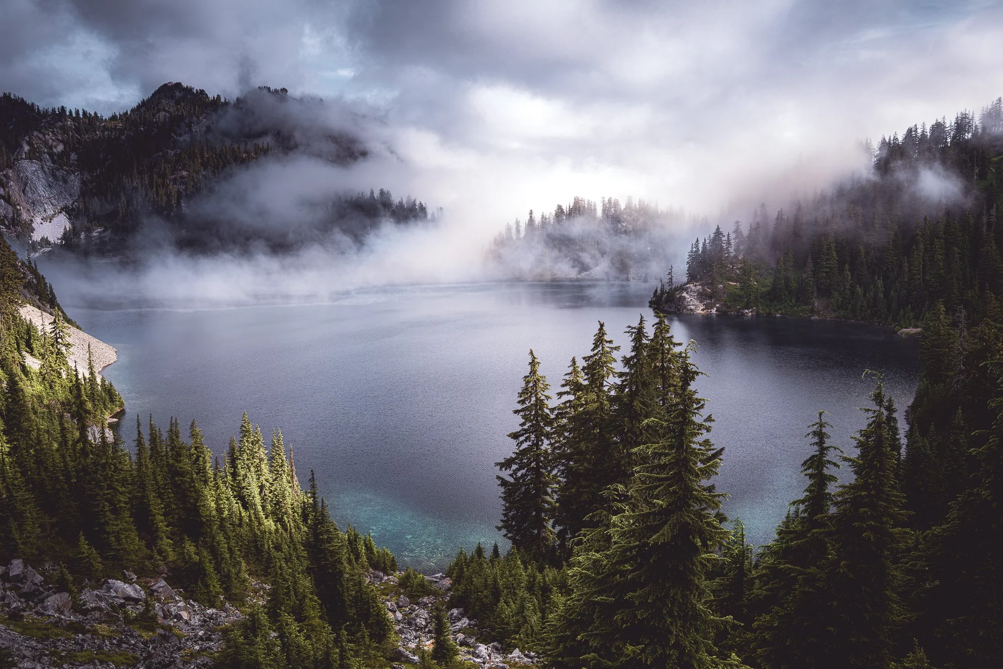 A mountain lake surrounded by dense evergreen trees, with fog covering the distant mountains and cloudy sky.