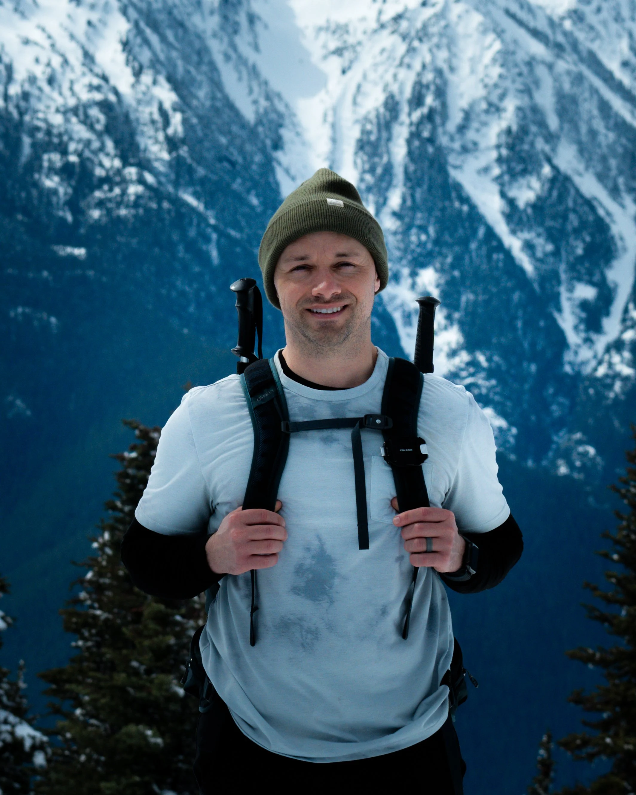 Man hiking in snowy mountainous terrain with evergreen trees, wearing a beanie and backpack.