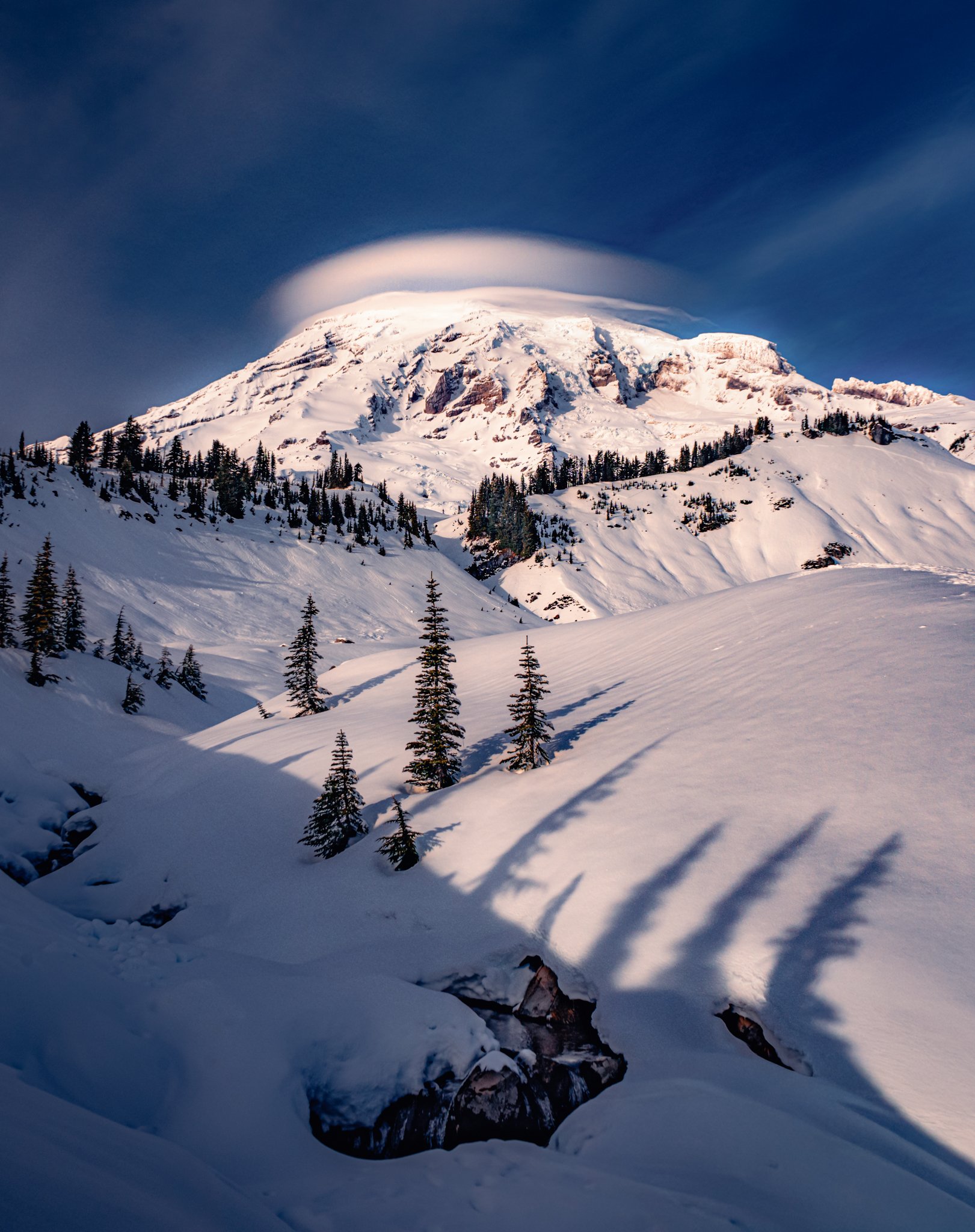 Snow-covered mountain with a cloud at the summit, surrounded by snow and pine trees in a winter landscape.