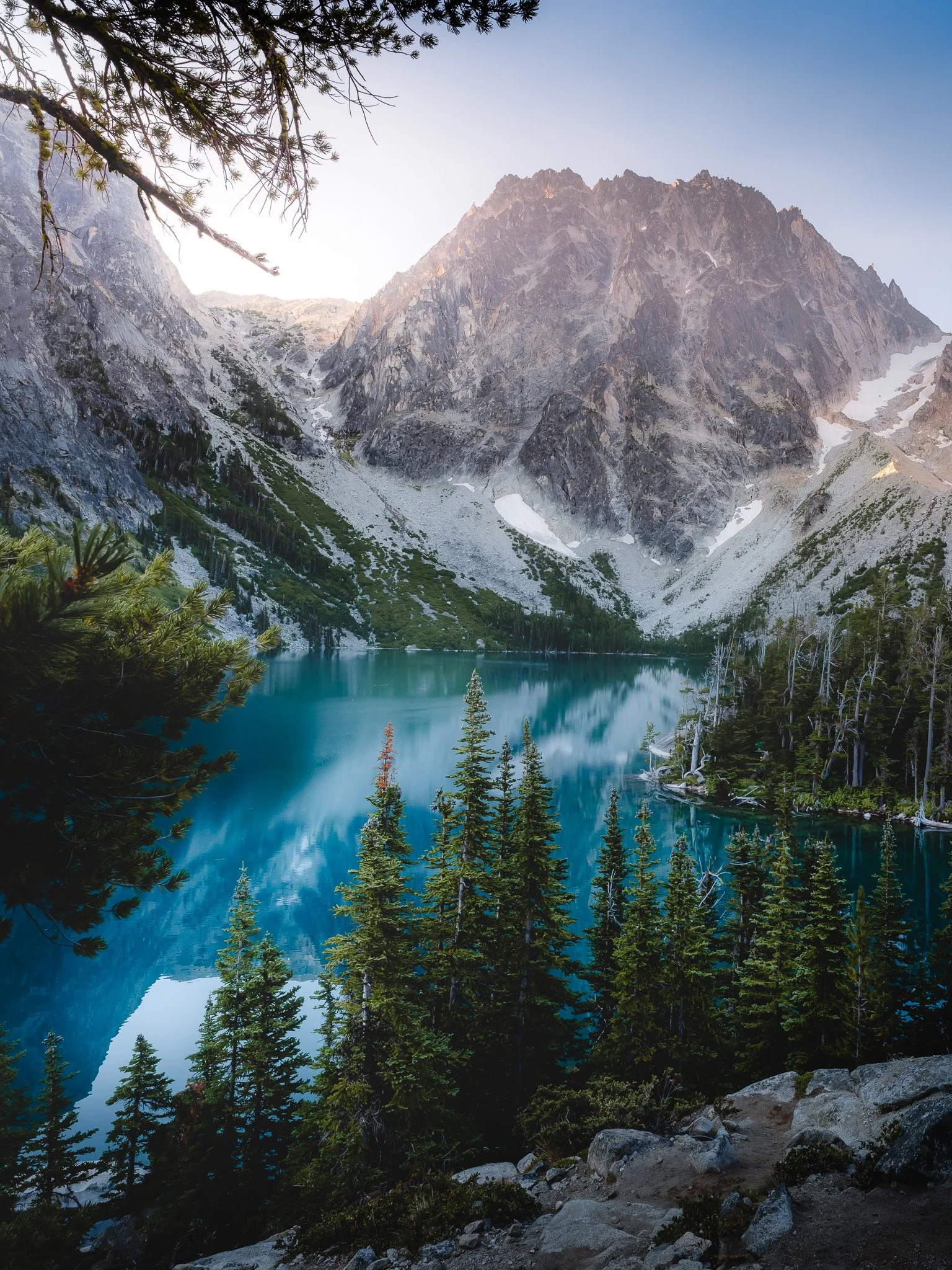 A scenic mountain landscape featuring a turquoise lake, evergreen trees in the foreground, and rugged mountains with patches of snow in the background.