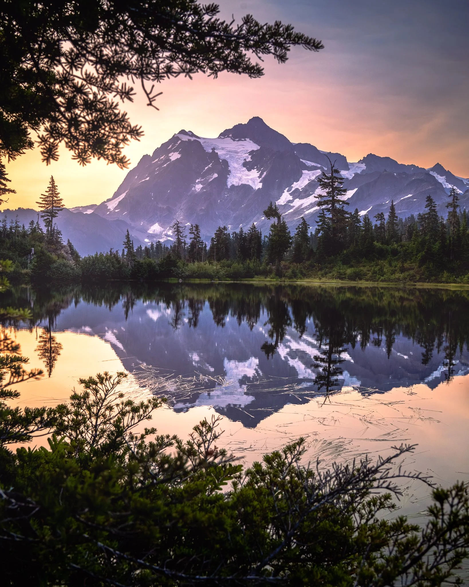 Snow-capped mountain reflecting in a calm lake at sunrise with trees in the foreground.