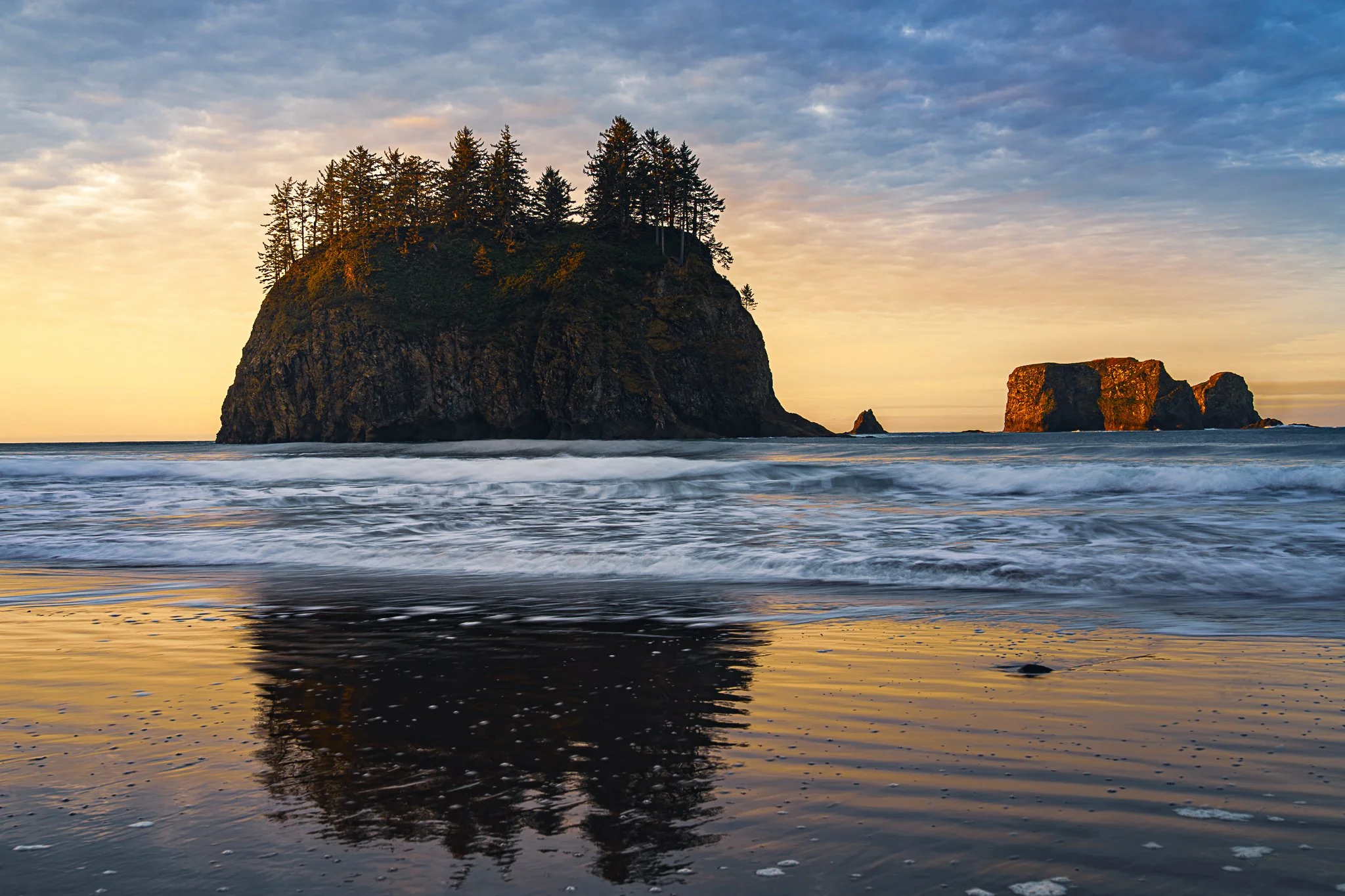 A large sea stack with trees on top, reflected in the wet sand of a beach at sunset, with a smaller rock formation in the distance and a cloudy sky overhead.