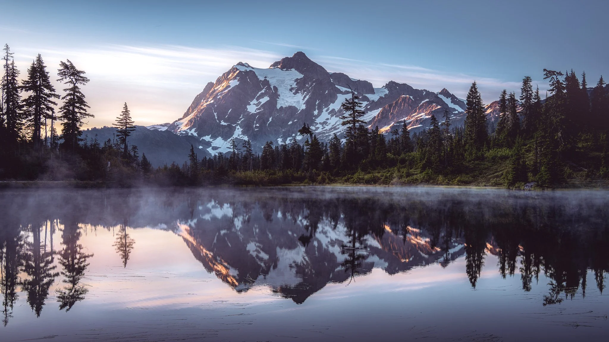 Snow-capped mountain peaks reflect in a still lake surrounded by pine trees and lush greenery, with a mist rising from the water and a partly cloudy sky overhead.