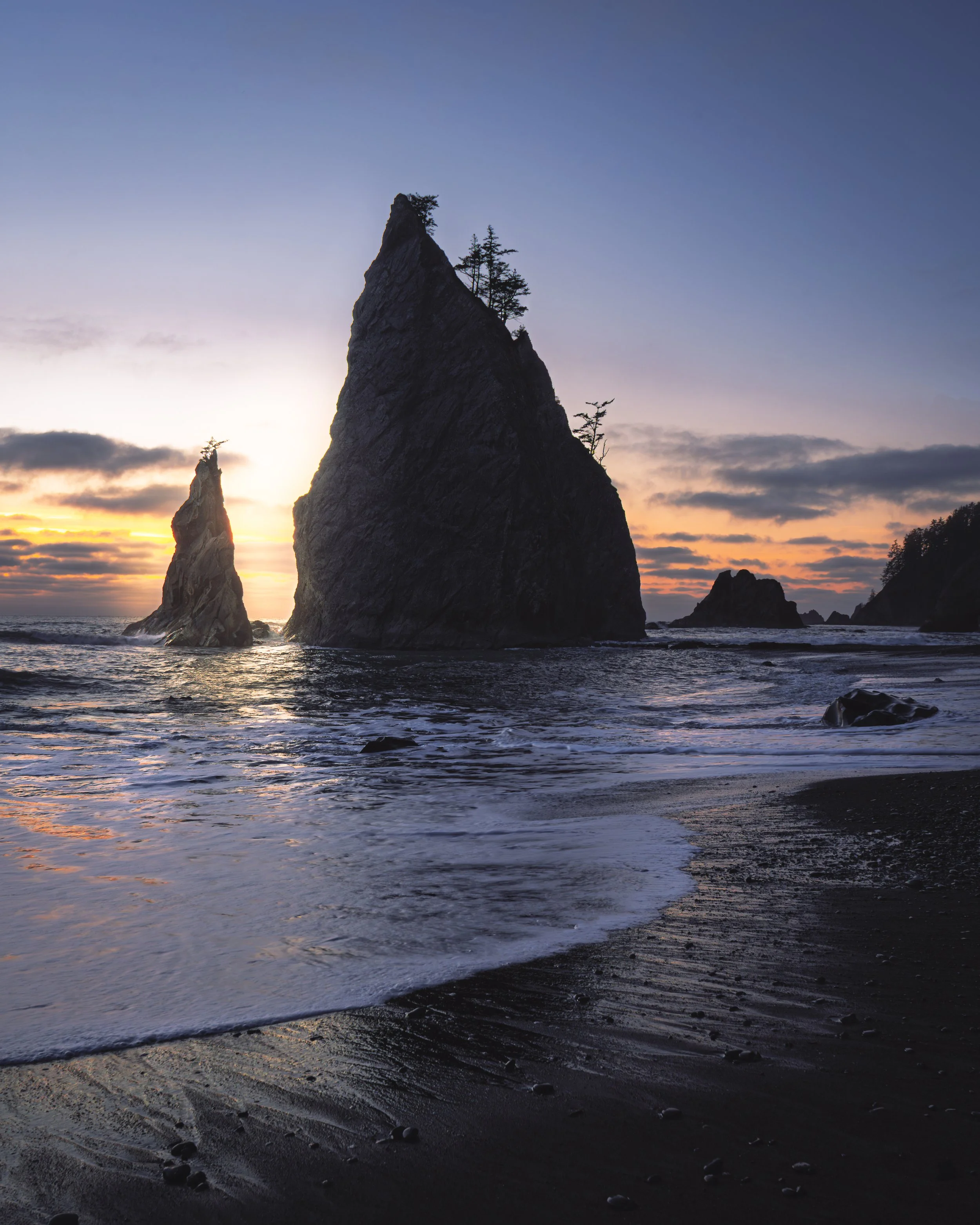 Sunset over rocky ocean with three large sea stacks and a few trees on top, waves crashing against black sandy beach.