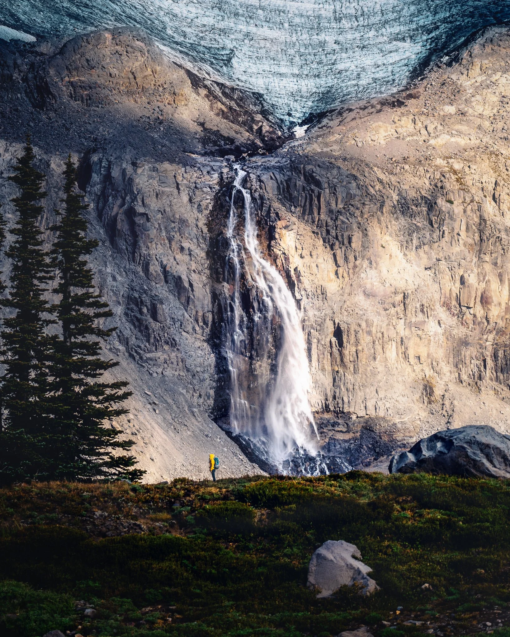 A hiker standing at the base of a large waterfall cascading down a rocky mountain face, surrounded by green vegetation.