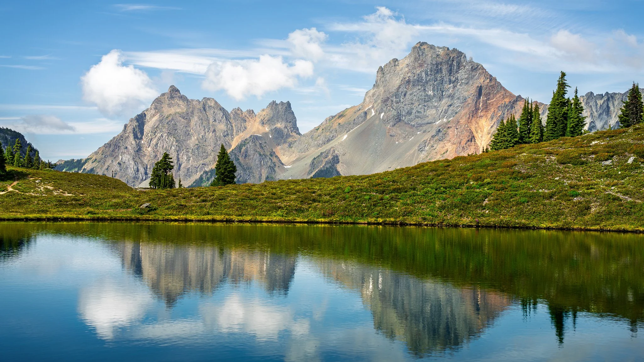 A mountain landscape with rocky peaks, a reflective lake in the foreground, grassy slopes, and scattered evergreen trees under a partly cloudy sky.