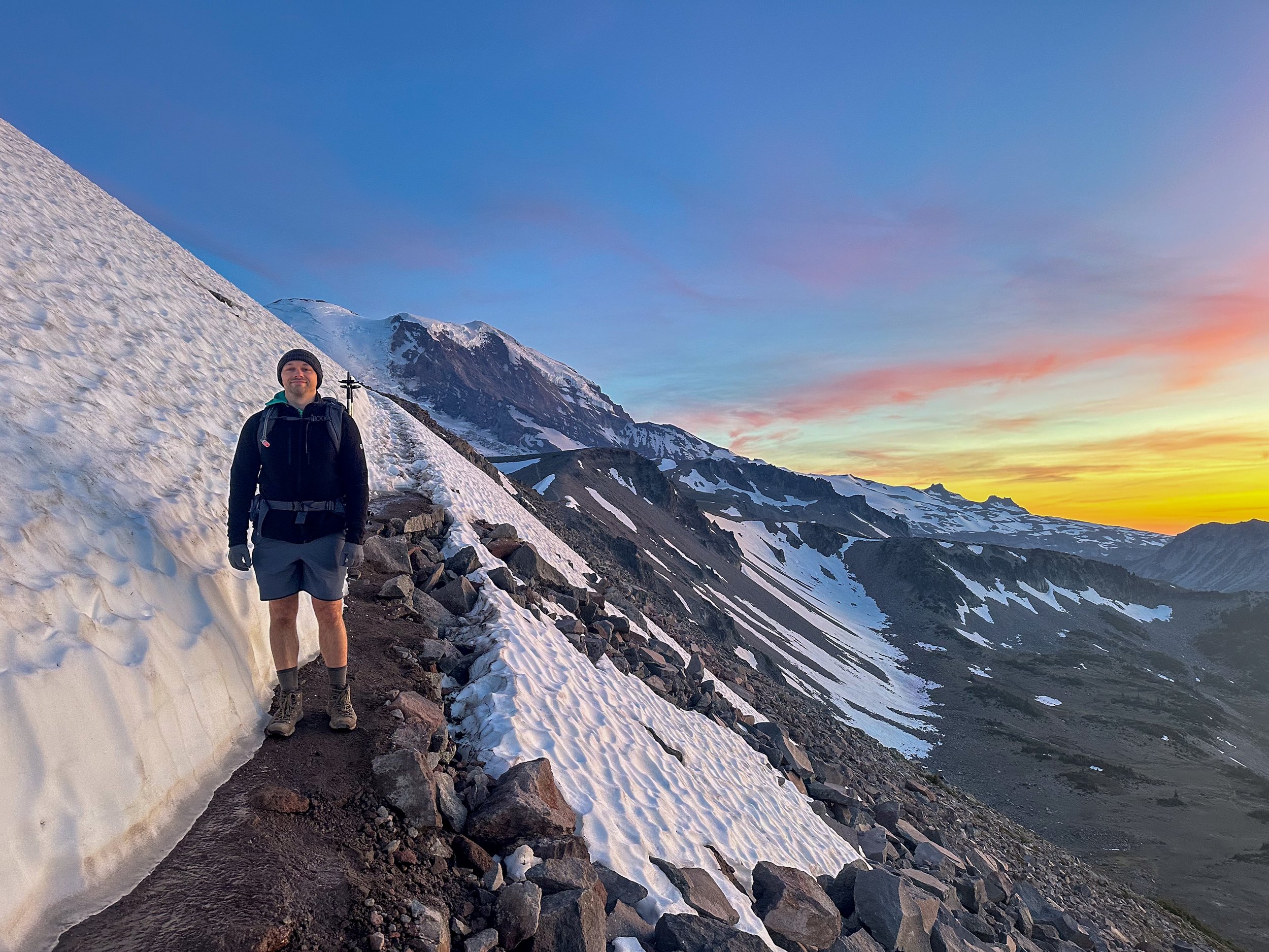 Man hiking on a snow-covered mountain trail with rugged terrain at sunset.