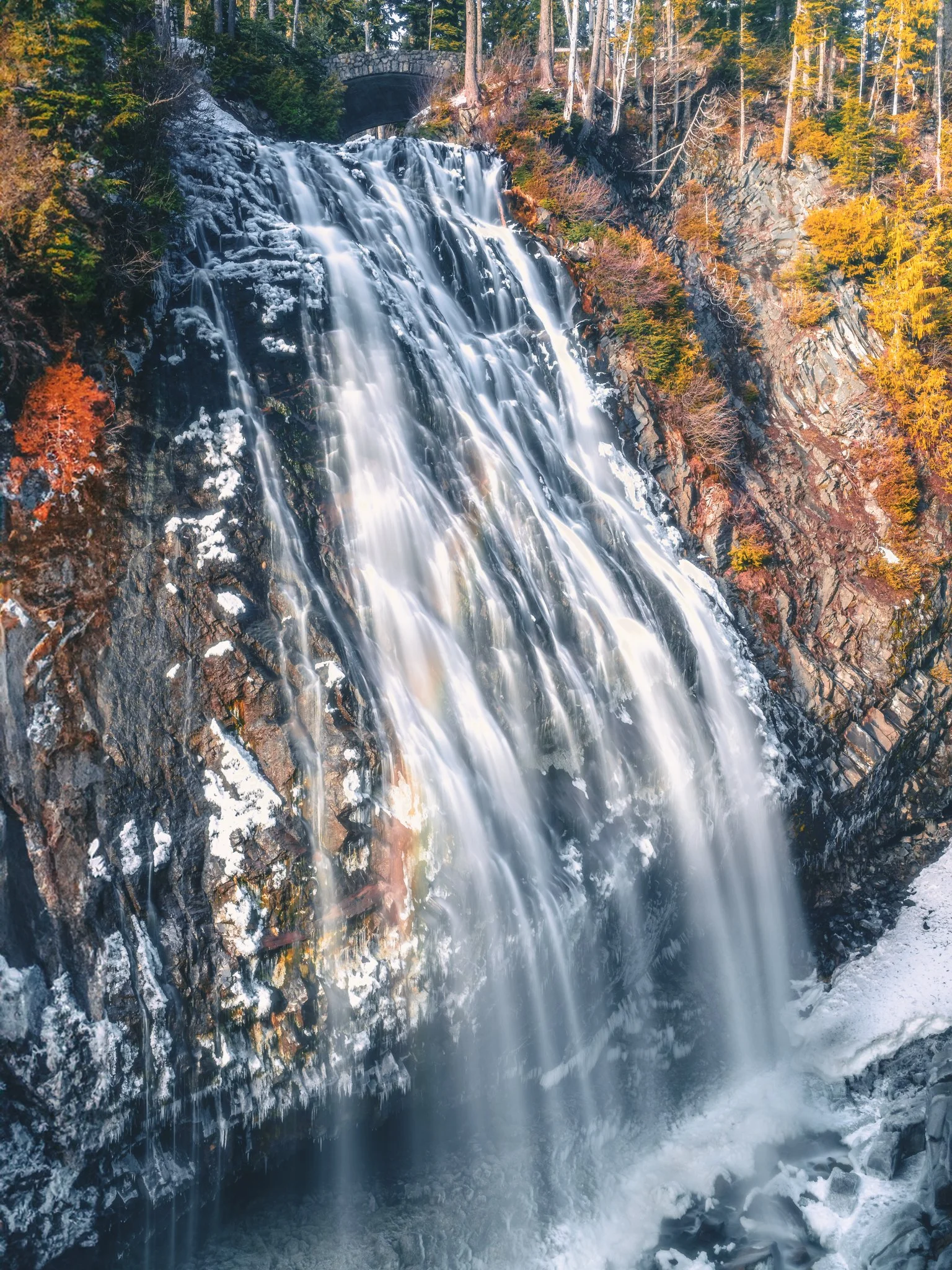 A waterfall flowing over rocks surrounded by autumn-colored trees, with a bridge visible at the top of the image.