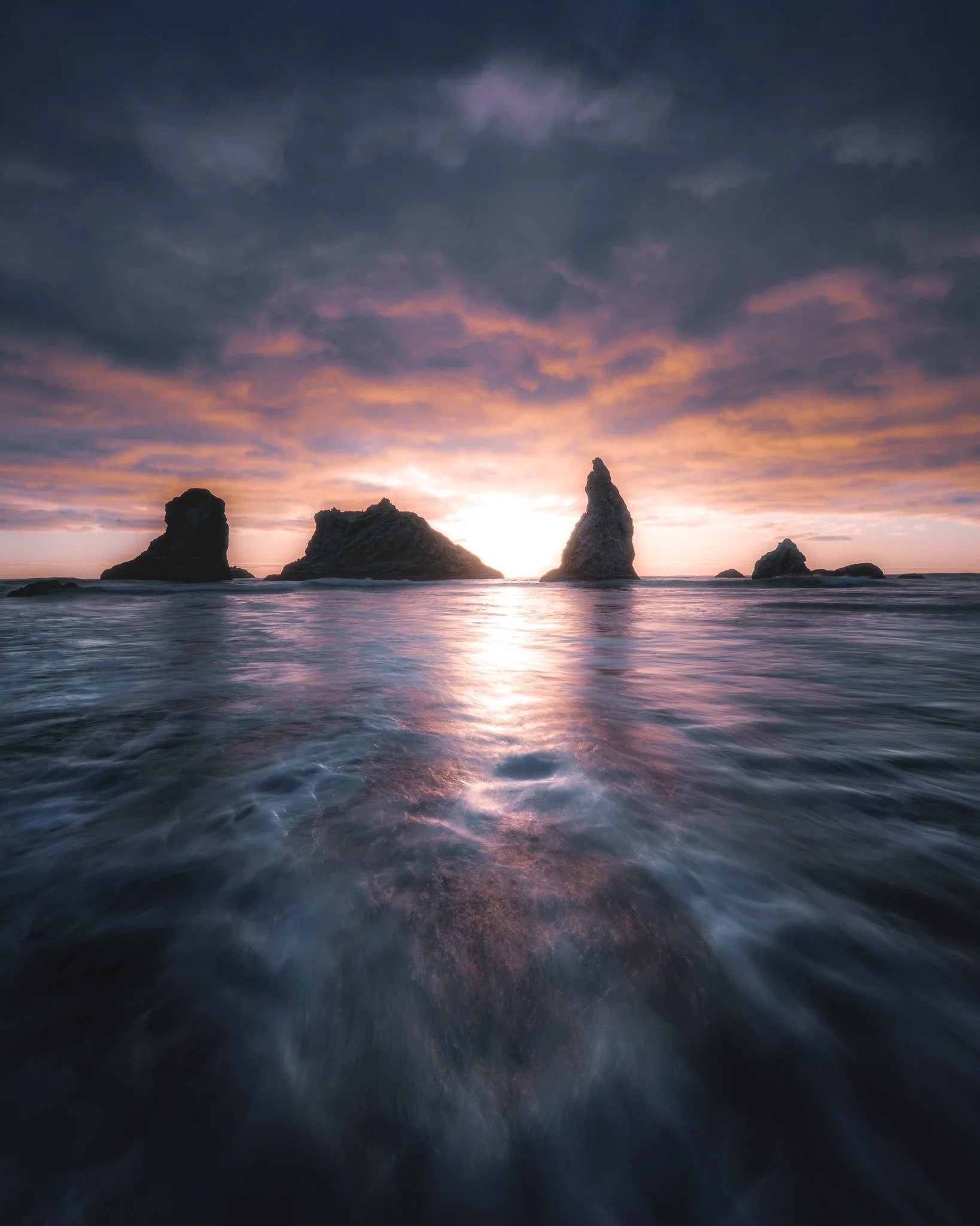 Sunset over the ocean with rocks protruding from the water and colorful sky with clouds.