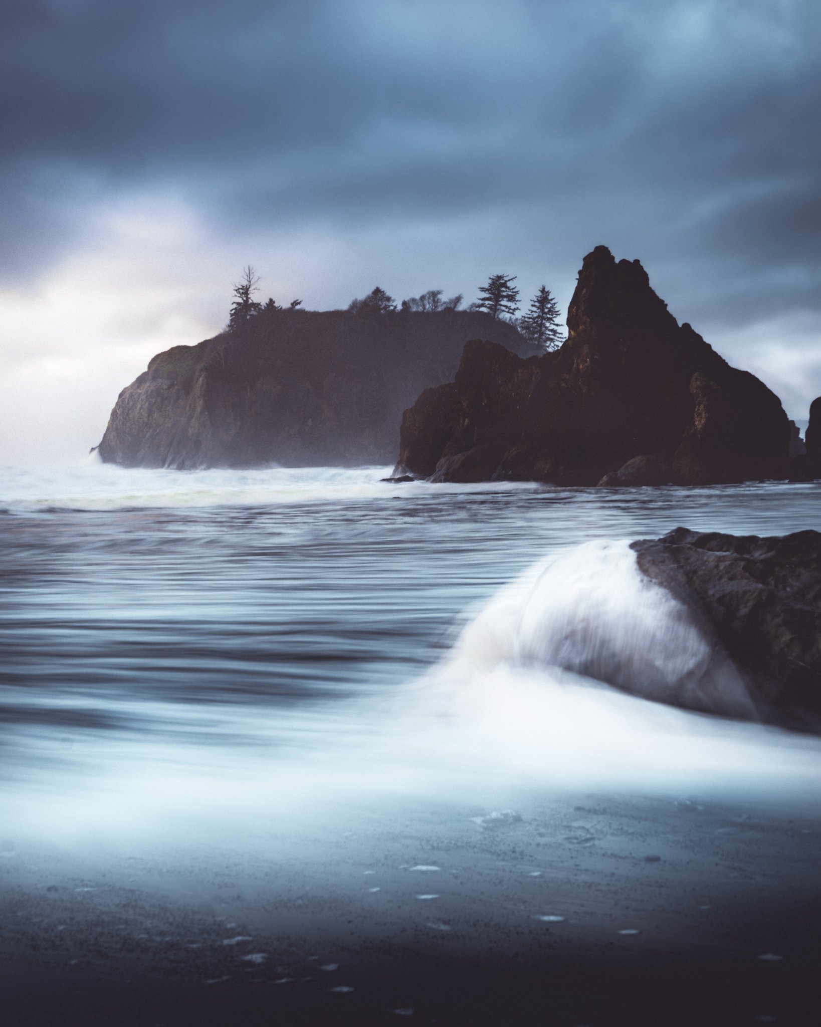 Coastal scene with large rock formations, trees on a cliff, and ocean waves under a cloudy sky