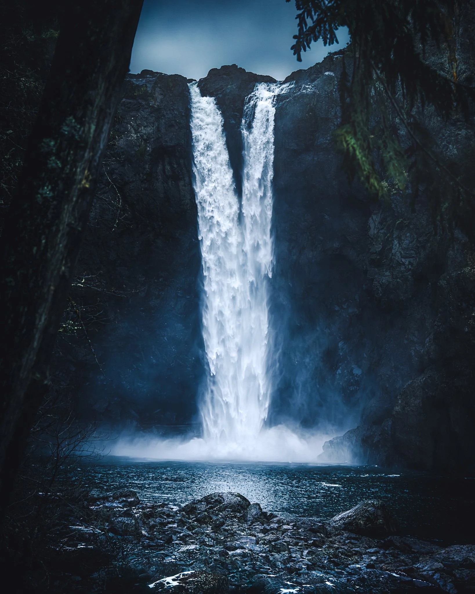 A tall waterfall cascading down a dark rocky cliff into a river, surrounded by trees.