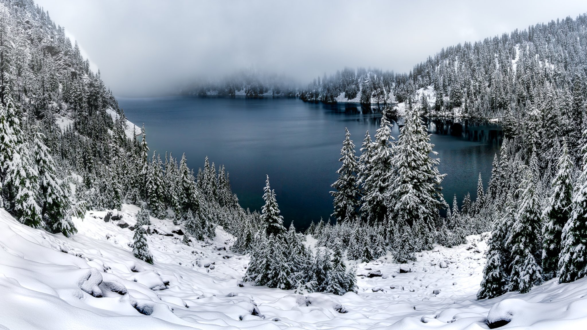 Snow-covered trees surrounding a mountain lake with foggy mountains in the background.
