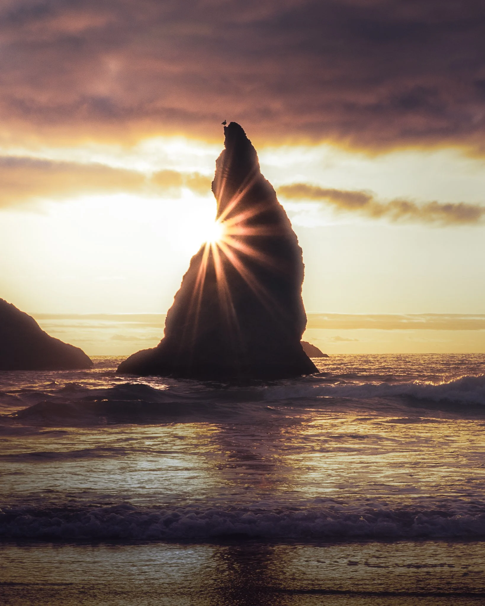 Sea stack rock formation at sunset with the sun shining through, creating a starburst effect, over an ocean with gentle waves.