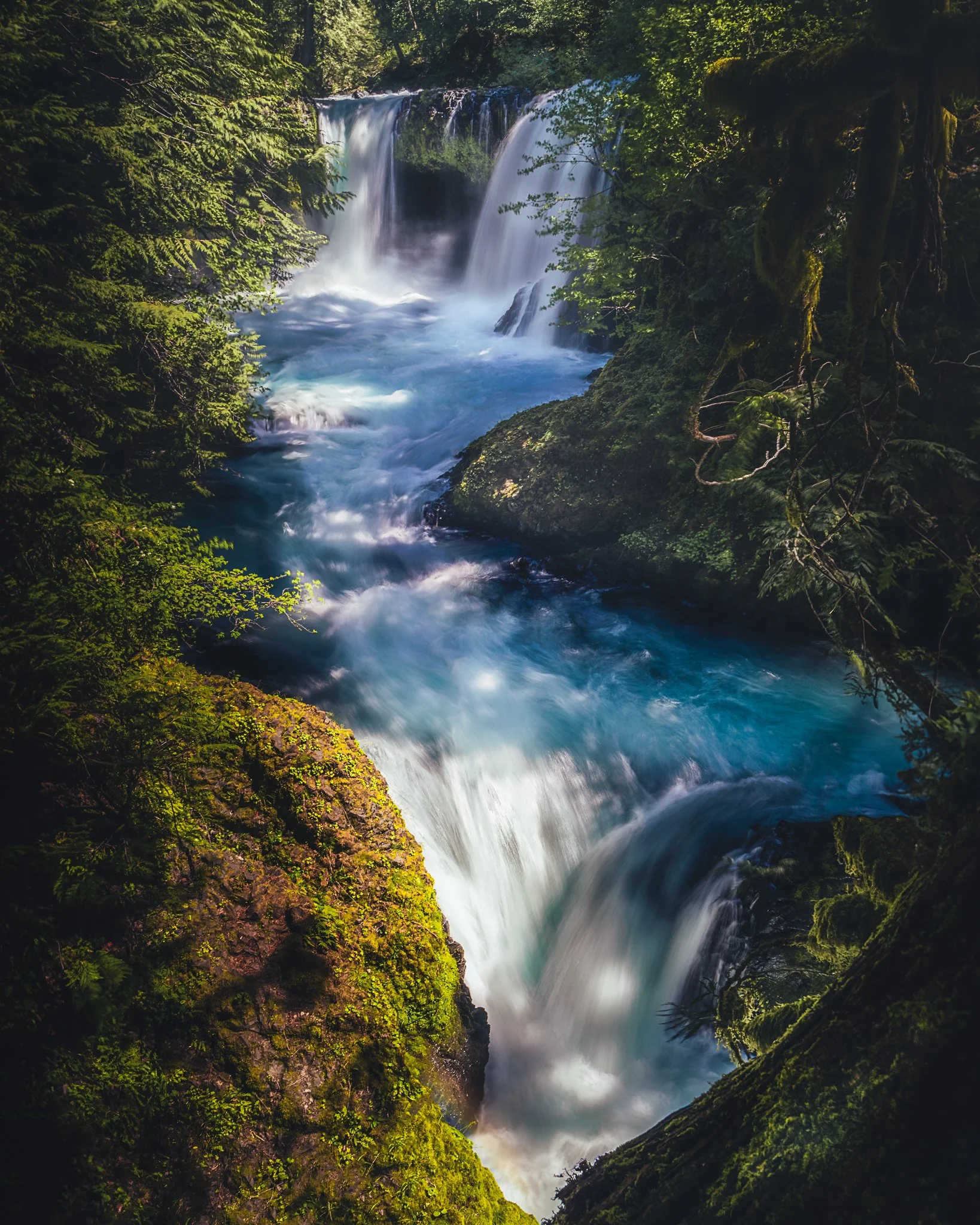 A scenic view of a multi-tiered waterfall flowing through a lush green forest with moss-covered rocks and vibrant foliage.
