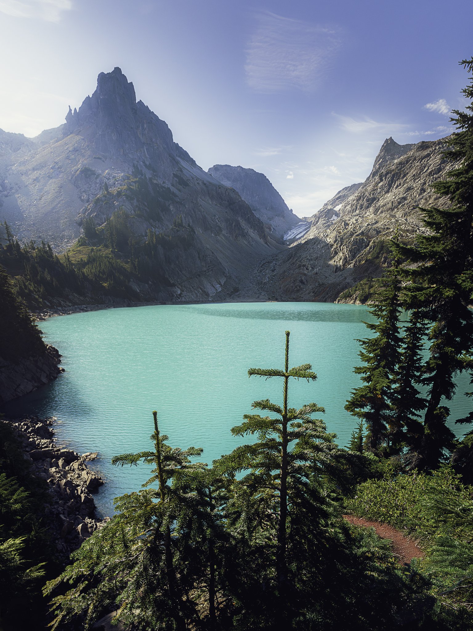 A scenic mountain landscape with a turquoise lake, surrounded by pine trees and rugged mountains under a blue sky.