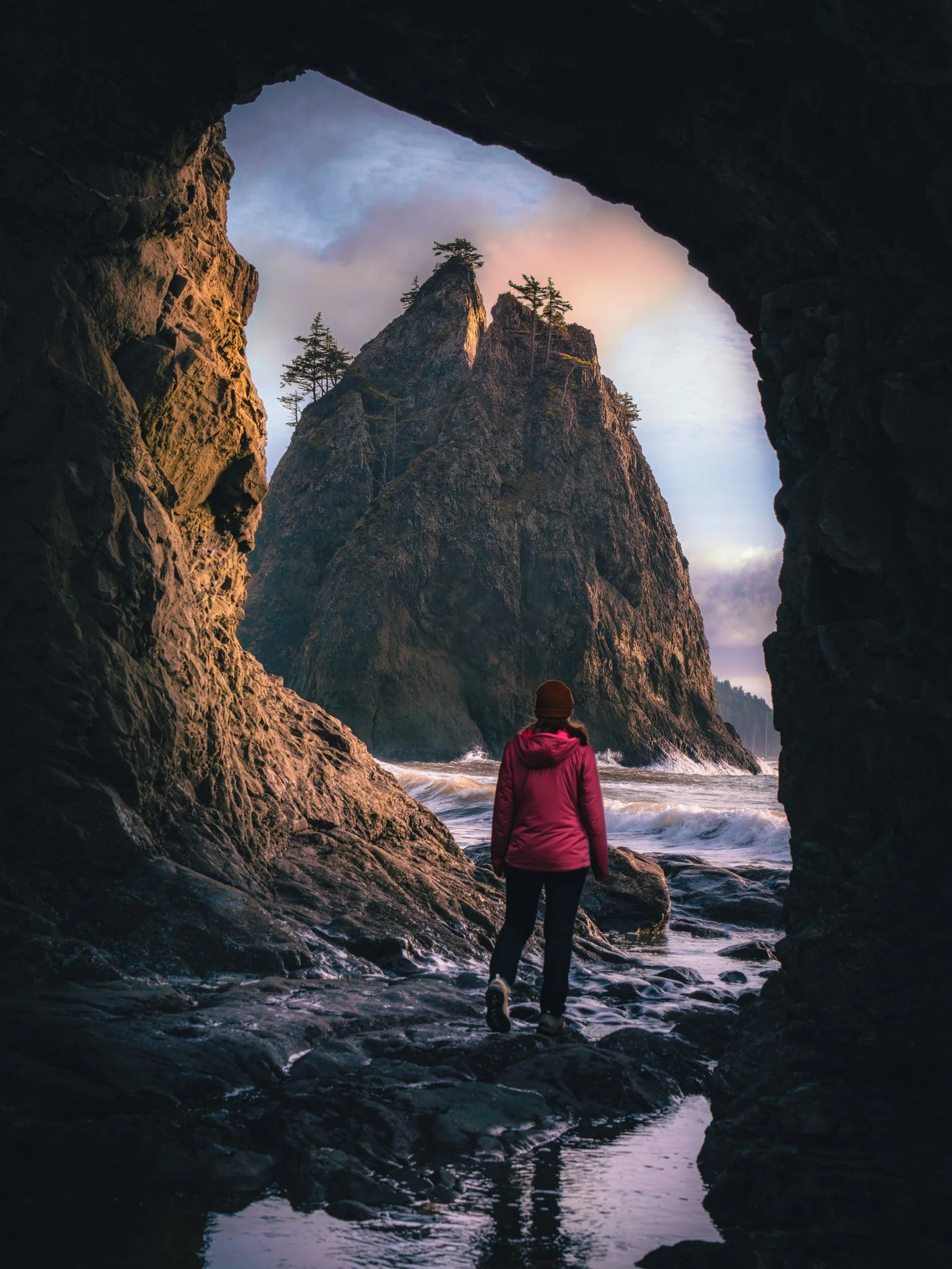 A person in a pink jacket and brown hat walking through a rocky coastal cave towards a large sea stack with trees on top, seen during sunset or sunrise.