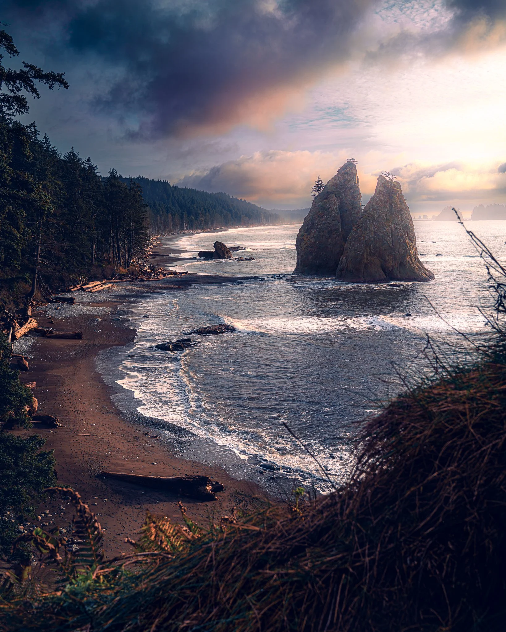 A scenic coastal landscape with two large sea stacks in the ocean, a forested shoreline, and a cloudy sky at sunset.