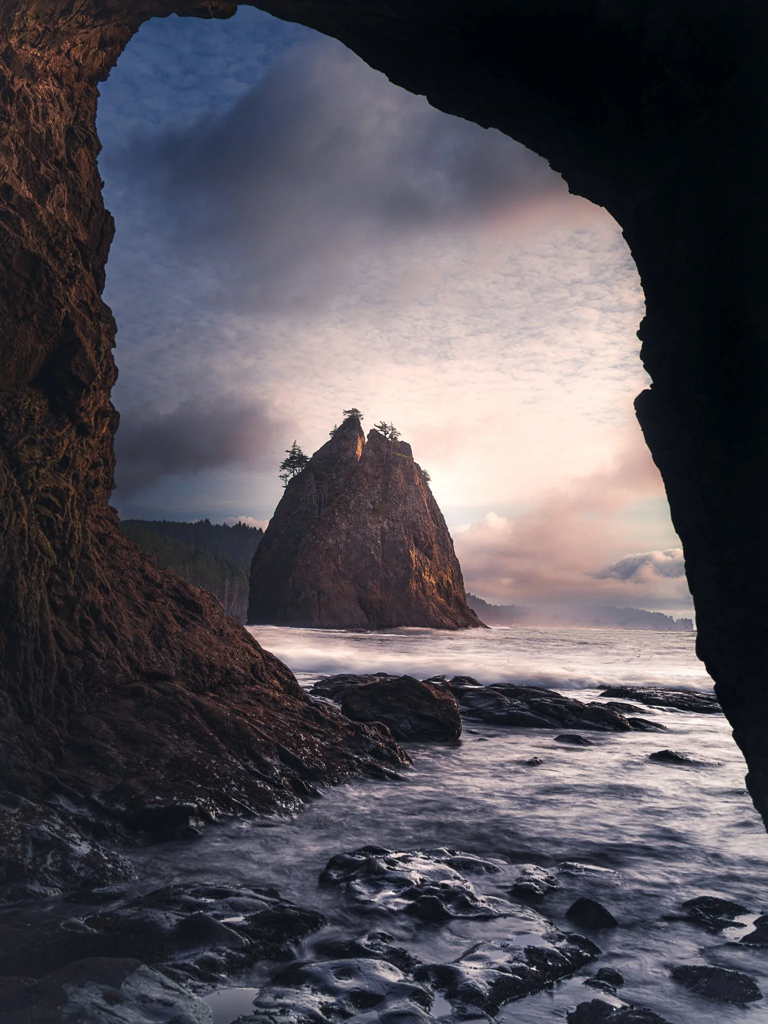 View of a sea stack seen through a rocky cave opening with a cloudy sky and waves crashing against rocks.