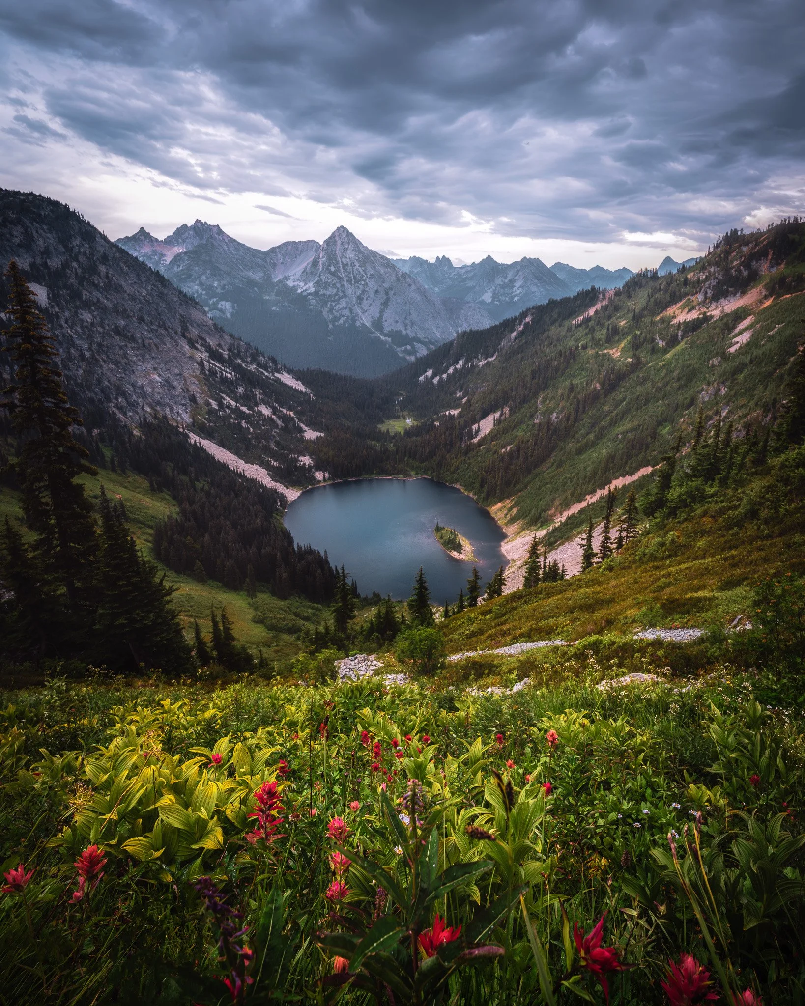 A scenic mountain landscape with a lake in the valley, surrounded by lush green hills, dense trees, and towering mountain peaks under a cloudy sky.
