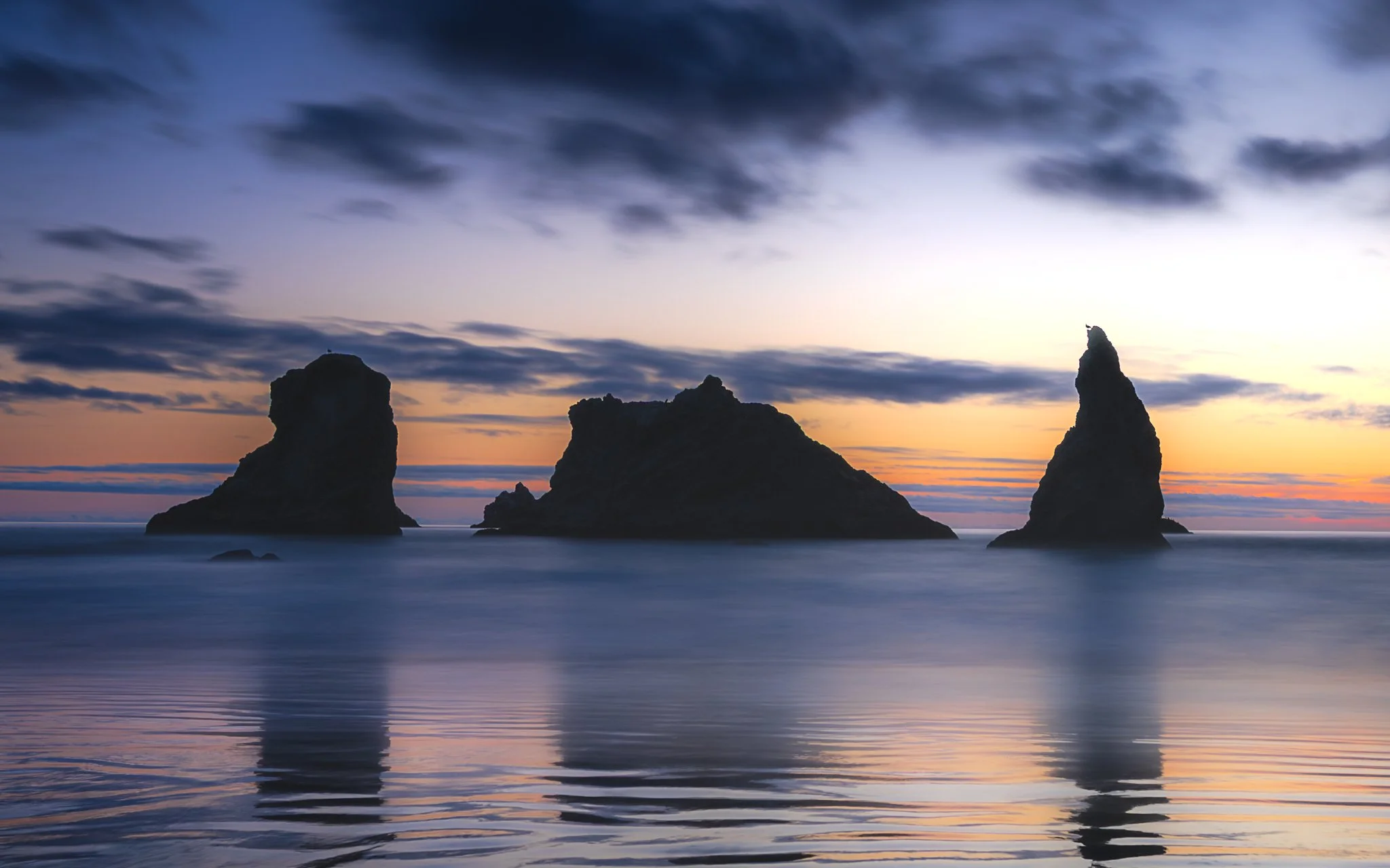 Sea stacks rising from calm ocean water during sunset, with a partly cloudy sky and pastel-colored horizon.