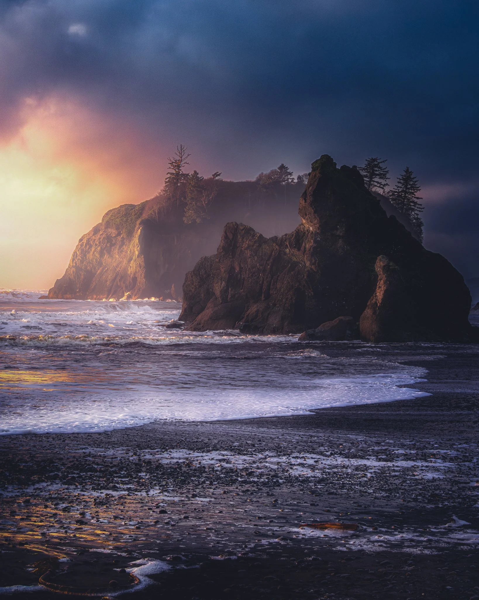 Sunset over rocky coastline with ocean waves, dark clouds, and a misty, forested cliff in the distance.